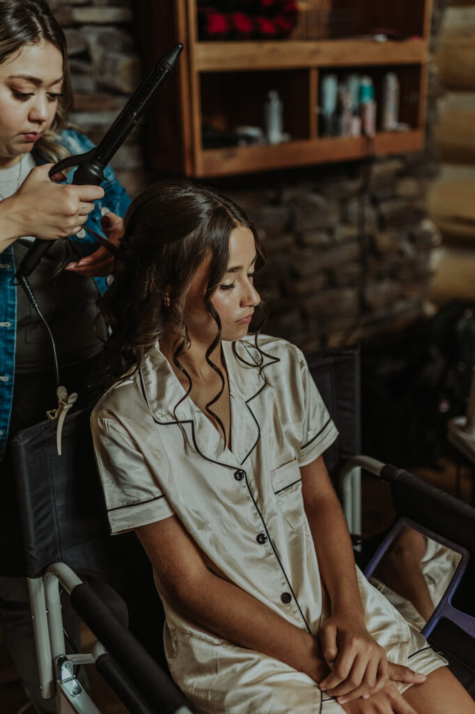 A wedding photo of a bridesmaid in silk pajamas having her hair curled by a hair stylist.