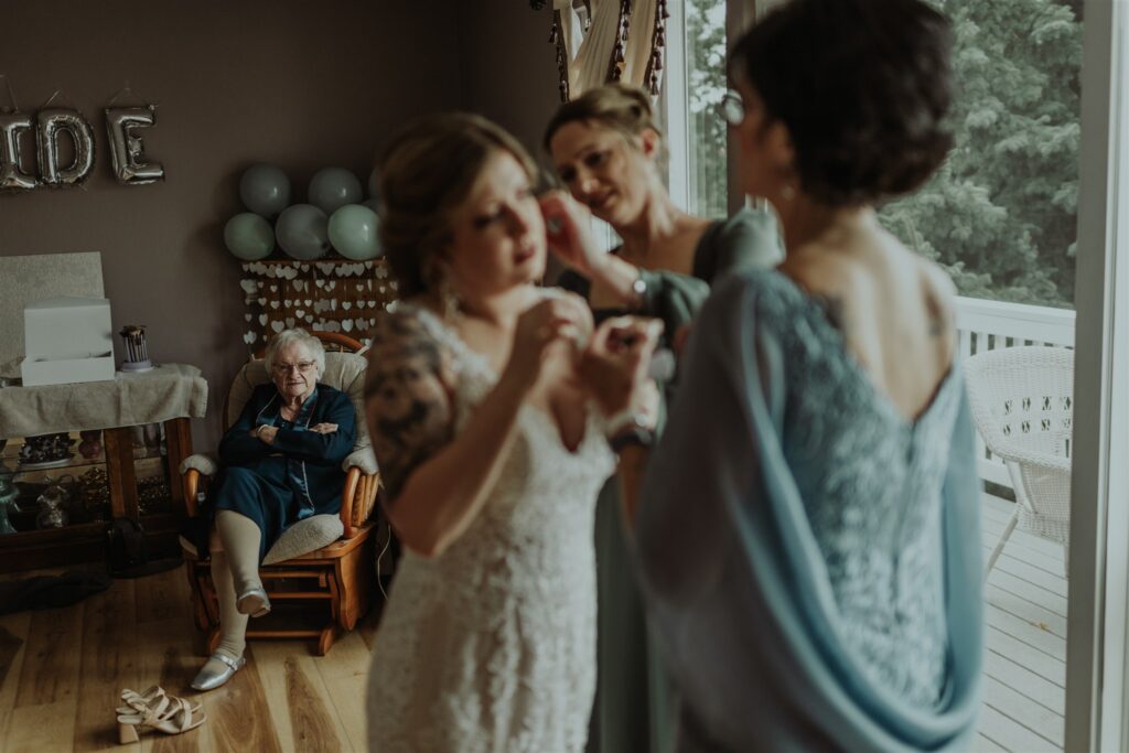 A documentary-style wedding photo of a bridesmaid puts a bride’s earring on as their mother watches on. In focus in the background is a grandmother watching them with love.