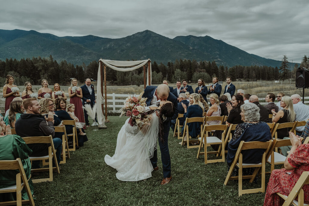 A wedding photo of a groom and bride kissing as they walk back down the aisle after their wedding ceremony. The groom is dipping the bride as their guests cheer. The ceremony is in the middle of a green valley surrounded by Montana mountains.