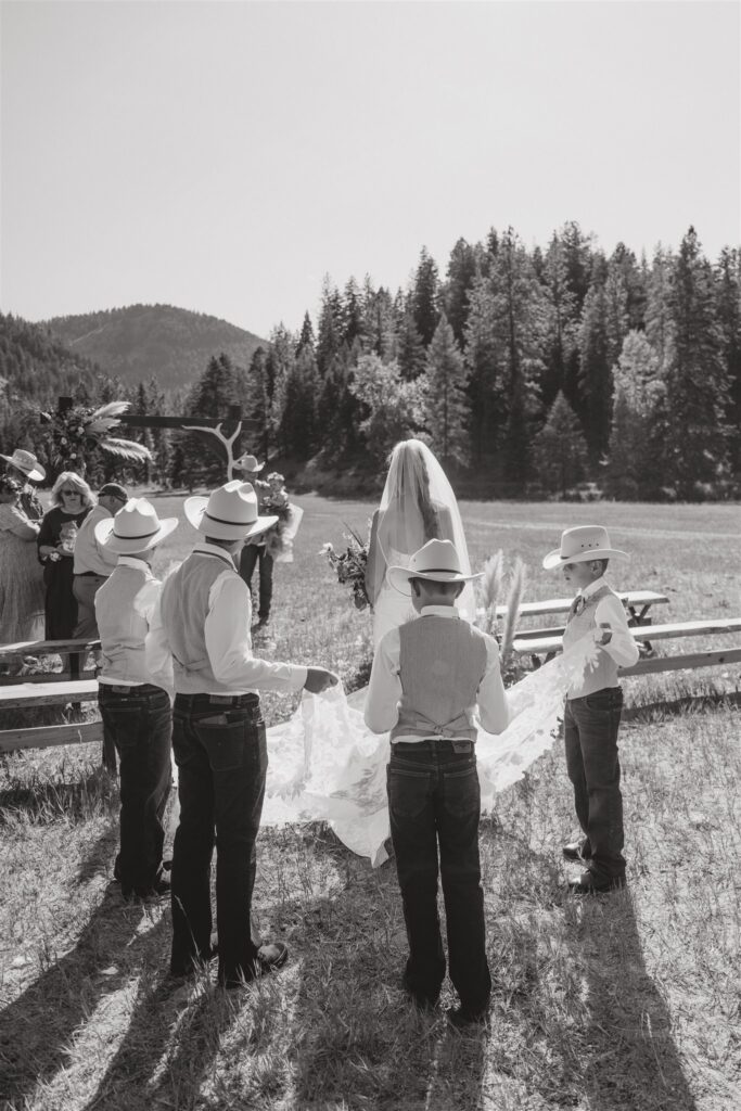 Black and white documentary-style wedding photography of a bride walking down the aisle during her ceremony. Her four sons are wearing cowboy hats and carrying her train.