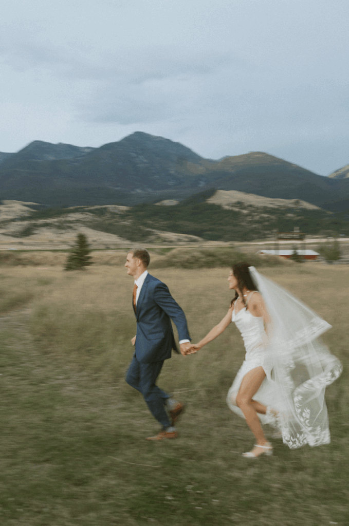 A photo by a Montana wedding photographer of a bride and groom running through a meadow with mountains in the background.