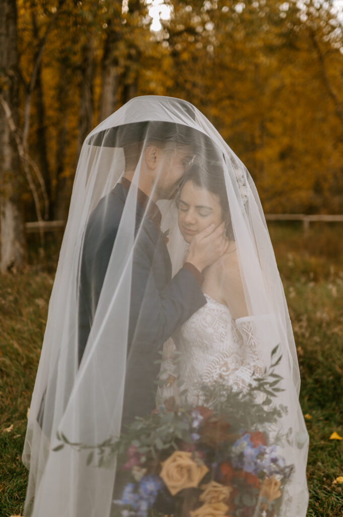 A wedding photo that feels authentic of a bride and groom almost kissing. There is a bridal veil over them and a forest in the background.