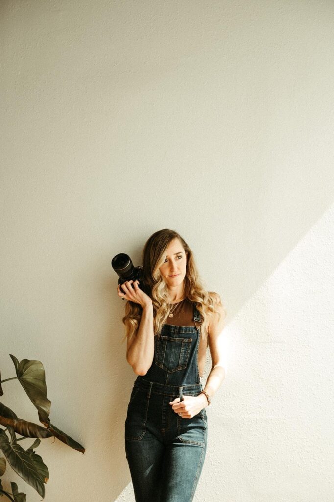 A photo of Montana wedding photographer Alicia Magnus posing with her camera wearing overalls, a burgundy tank top, and a white cardigan.